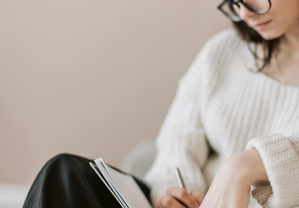 Attentive female wearing eyeglasses and casual outfit sitting barefoot with crossed legs on comfortable couch in modern flat and taking notes in notebook with pen